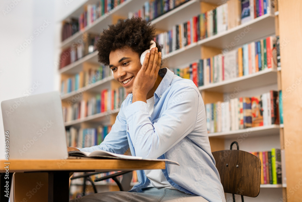 Positive Latin student guy studying in college library, using laptop and learning online for university exam, wearing headphones and smiling Positive Latin student guy studying in college library, using laptop and learning online for university exam, wearing headphones and smiling