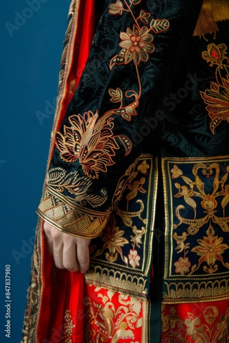 Close-up of a person wearing an intricate, ornate embroidered traditional garment with floral patterns and red and black fabric on a blue background.
