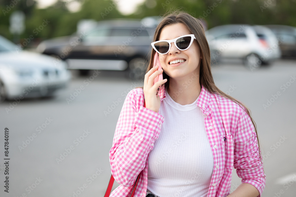 using the phone. telephone conversation. positive smile woman wearing white vichy cage t-shirt and pink checkered shirt sunglasses smiles while talking her phone. Standing city street blurred parking using the phone. telephone conversation. positive smile woman wearing white vichy cage t-shirt and pink checkered shirt sunglasses smiles while talking her phone. Standing city street blurred parking