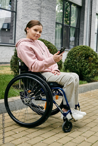 A young woman in a pink hoodie sits in a wheelchair outside, smiling as she uses her phone. photo