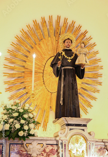 Statue of saint anthony of padua holding baby jesus and a lily, standing inside a church in front of a golden halo