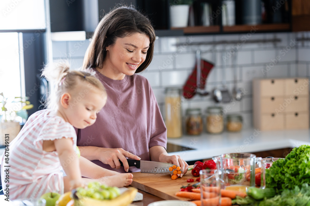 A cheerful mother is making a healthy meal with lots of fruit and vegetables, and her cute little baby daughter is keeping her company