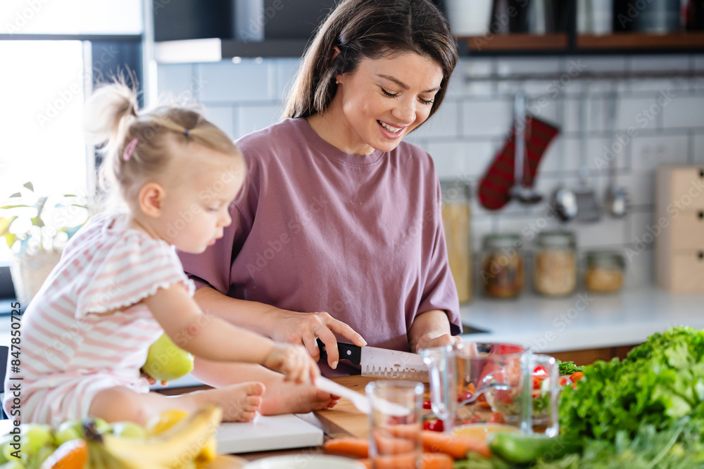A cheerful mother is making a healthy meal with lots of fruit and vegetables, and her cute little baby daughter is keeping her company A cheerful mother is making a healthy meal with lots of fruit and vegetables, and her cute little baby daughter is keeping her company