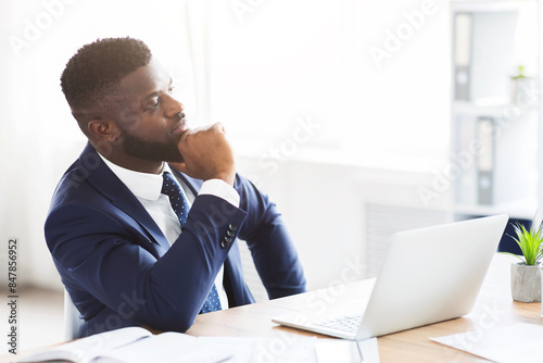 Thoughtful afro businessman making business solution at workplace, touching chin and looking aside, empty space