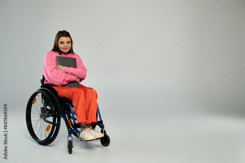 A young brunette woman in casual attire sits in a wheelchair in a studio setting, holding a laptop in her lap. A young brunette woman in casual attire sits in a wheelchair in a studio setting, holding a laptop in her lap.