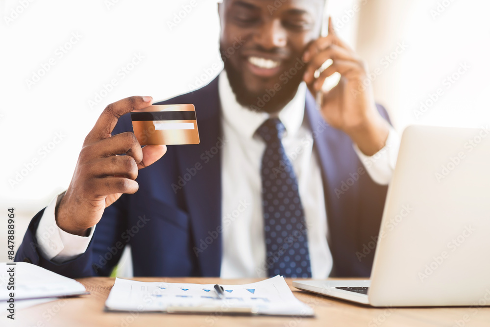 Easy payments. Happy african manager holding credit card after online purchase and talking on mobile phone with customer service, cropped Easy payments. Happy african manager holding credit card after online purchase and talking on mobile phone with customer service, cropped