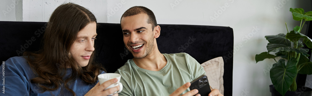 A young gay couple relaxes in bed, sharing a quiet moment together. One partner holds a cup, while the other looks at a smartphone. A young gay couple relaxes in bed, sharing a quiet moment together. One partner holds a cup, while the other looks at a smartphone.