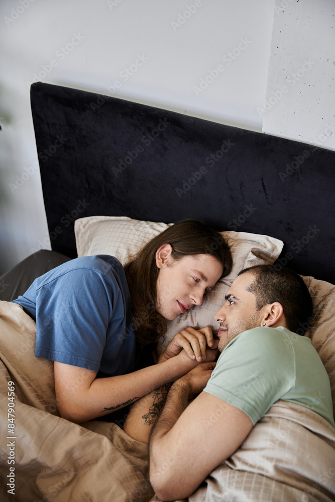 A young gay couple cuddles in bed, their eyes locked in a tender gaze, reflecting their deep love and connection. A young gay couple cuddles in bed, their eyes locked in a tender gaze, reflecting their deep love and connection.