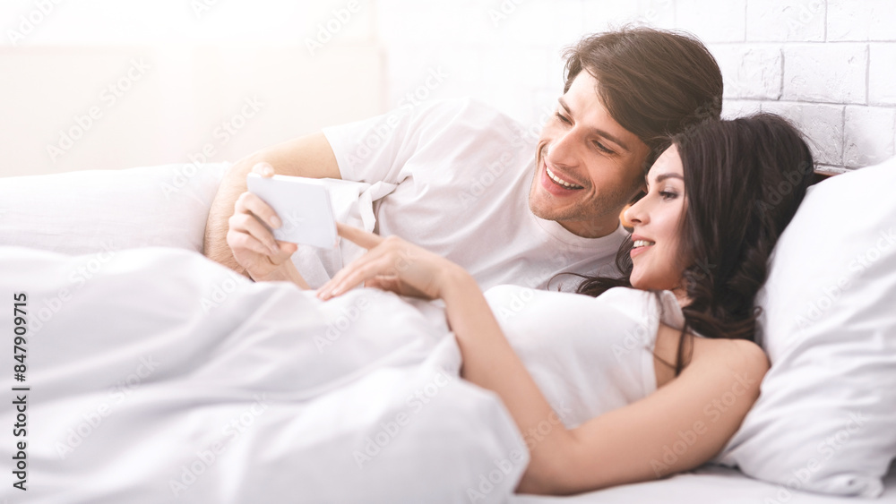 Fototapeta premium A young couple lies in bed together, covered in white sheets. The man is on his side, smiling and looking at the smartphone held in the woman hand.