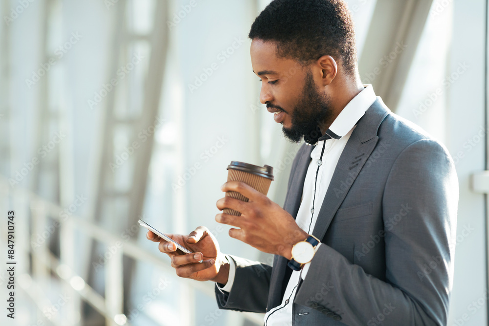 Smiling African American businessman holding a coffee cup while checking his smartphone in a bright, modern airport, free space Smiling African American businessman holding a coffee cup while checking his smartphone in a bright, modern airport, free space
