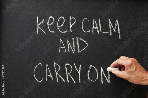 Hand with chalk writing quote Keep Calm And Carry On on blackboard