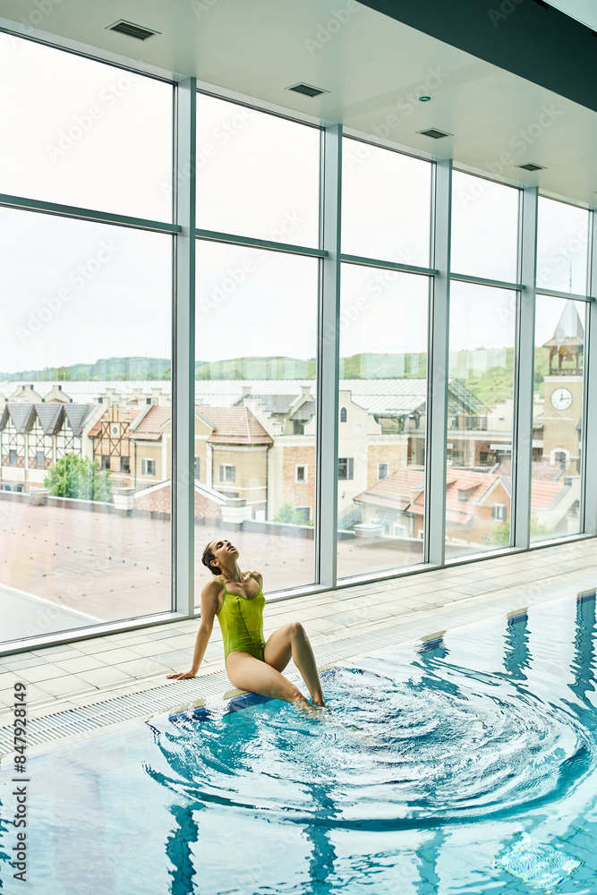 A young brunette woman in a green bathing suit sits gracefully on the edge of an indoor swimming pool. A young brunette woman in a green bathing suit sits gracefully on the edge of an indoor swimming pool.