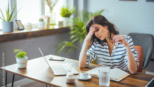 A thoughtful Caucasian businesswoman absently holds her glasses while reviewing documents beside her laptop, surrounded by office plants and a coffee cup, signifying a busy work environment. photo