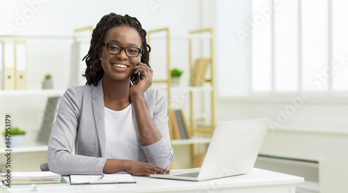 Happy young African American woman in formal attire, a manager using a laptop at the office, talking on the phone.