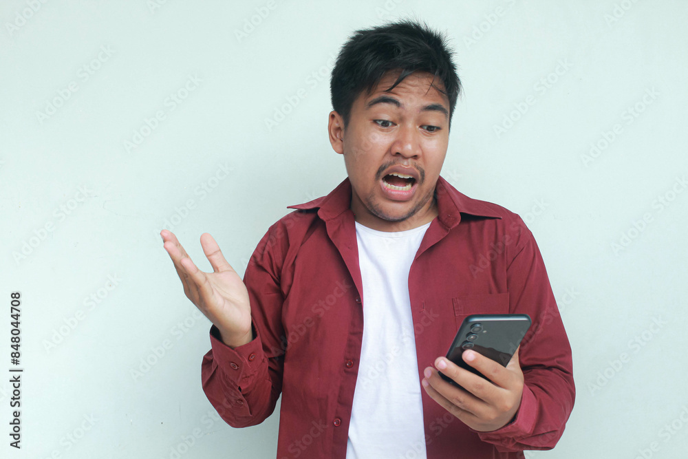 Portrait of unhappy and stressed Asian man in red shirt holding mobile phone with sad expression on face while scratching forehead. Isolated image on gray background. Portrait of unhappy and stressed Asian man in red shirt holding mobile phone with sad expression on face while scratching forehead. Isolated image on gray background.