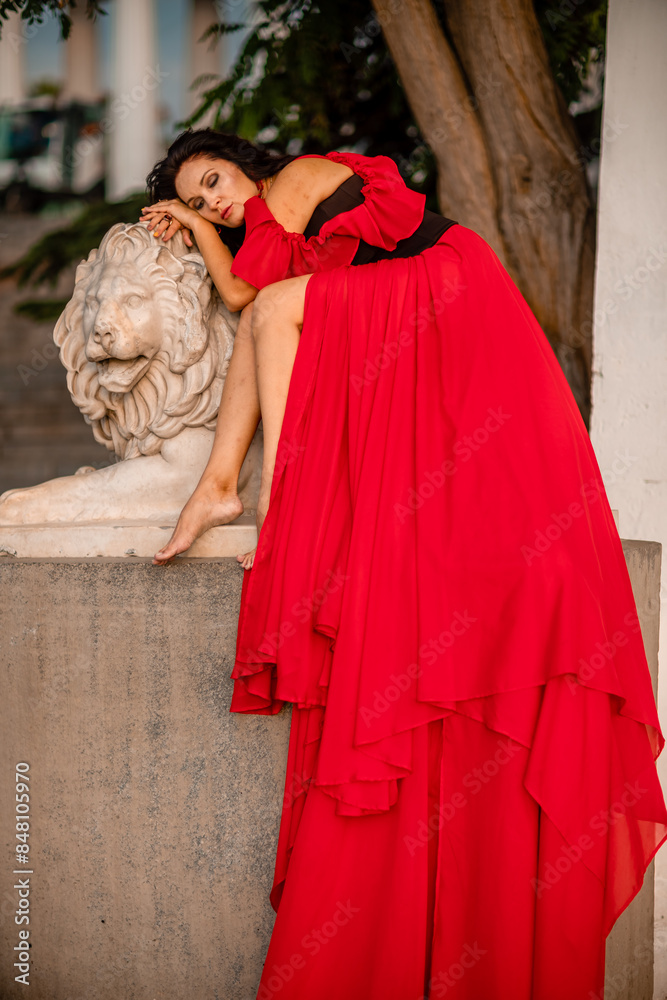 A woman in a red dress is sitting on a stone lion statue. The statue is white and has a lion's head. The woman is wearing a black belt and is looking at the camera. The scene has a calm. A woman in a red dress is sitting on a stone lion statue. The statue is white and has a lion's head. The woman is wearing a black belt and is looking at the camera. The scene has a calm.