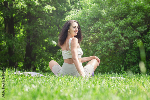 A young woman sits on a mat in a park on a sunny day. Beautiful smiling brunette in beige sportswear. Health and active lifestyle.