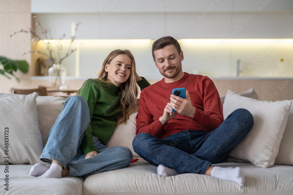 Contented married couple relaxing on couch at home, man shows wife smartphone screen, plan trip together, online shopping using phone. Cheerful husband show to woman fun video in social media on sofa. Contented married couple relaxing on couch at home, man shows wife smartphone screen, plan trip together, online shopping using phone. Cheerful husband show to woman fun video in social media on sofa.