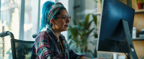 Inclusive photo of female employee with blue hair sitting in a wheelchair in diverse & accessible dei workplace. Happy remote worker at computer. Pride month business marketing photo