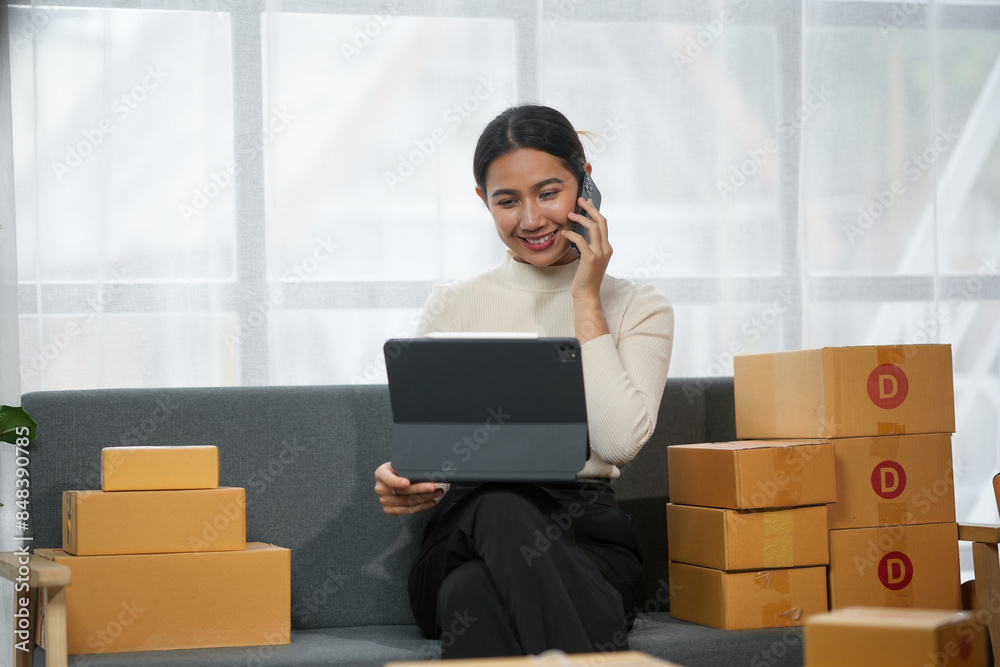 Smiling woman working on laptop while talking on phone, surrounded by cardboard boxes, indicating an online business or e-commerce activity. Smiling woman working on laptop while talking on phone, surrounded by cardboard boxes, indicating an online business or e-commerce activity.