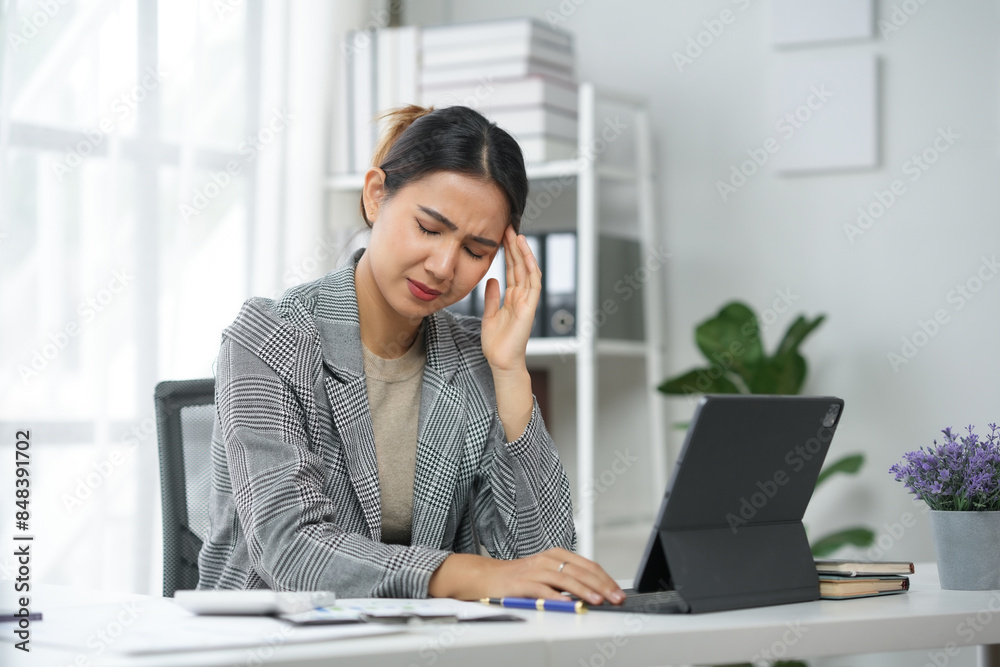 Woman in gray blazer sitting at desk with laptop, holding head in pain, experiencing stress or headache in modern office environment. Woman in gray blazer sitting at desk with laptop, holding head in pain, experiencing stress or headache in modern office environment.