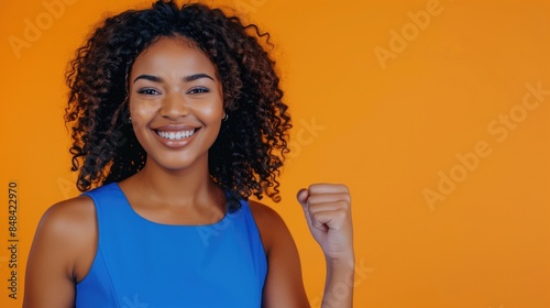 Beautiful young black woman in a blue dress before a solid orange colored background. She is in the side of the image, and and show her fist. photo