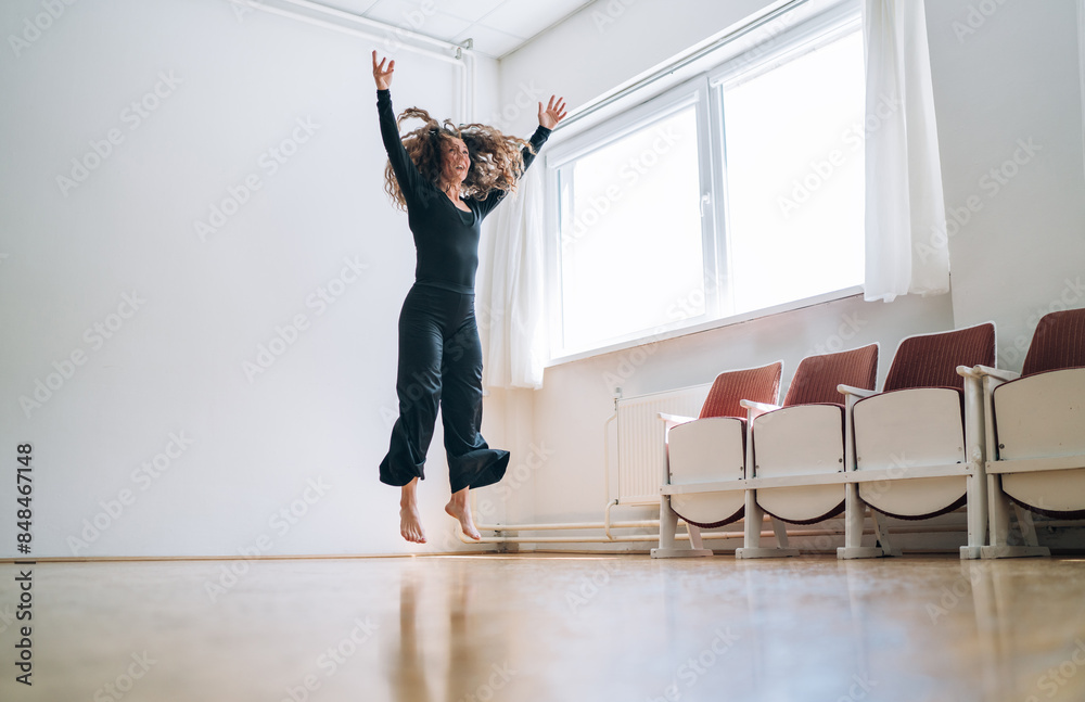 Fototapeta premium Woman in a black outfit gracefully jumping in a well-lit room, her movements fluid and expressive. Her curly hair flows as she extends her arms, embodying elegance and passion in her dance