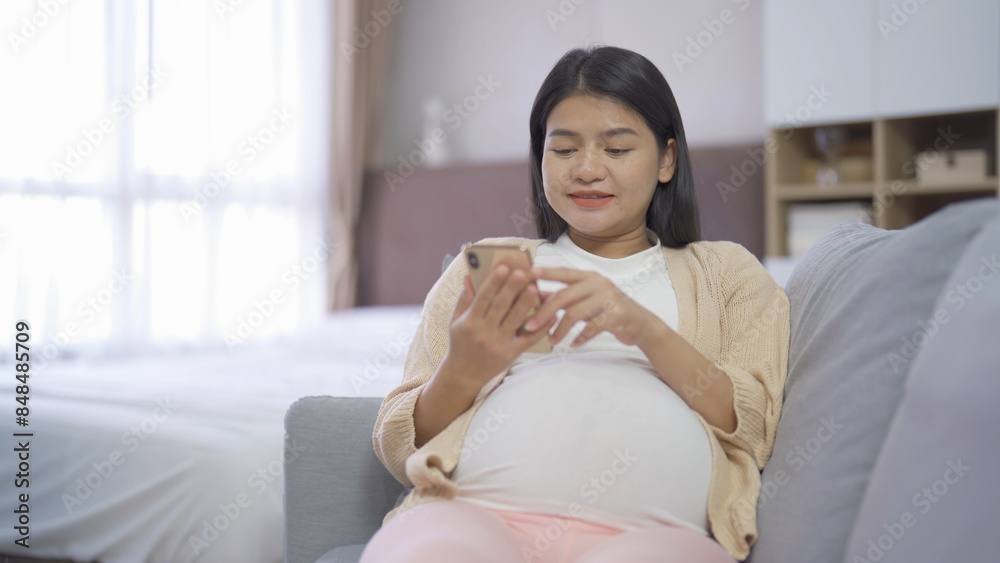 A portrait of happy Asian pregnant woman using online mobile phone technology device with screen in bedroom at home. Having a baby. Family people lifestyle. Mom love. A portrait of happy Asian pregnant woman using online mobile phone technology device with screen in bedroom at home. Having a baby. Family people lifestyle. Mom love.