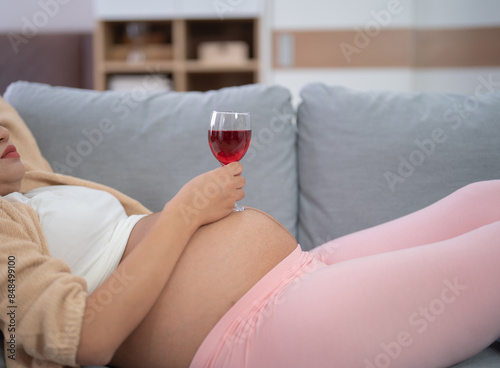 A portrait of happy Asian pregnant woman drinking wine alcohol beverage glass in bedroom at home. Having a baby. Family people lifestyle. Mom.