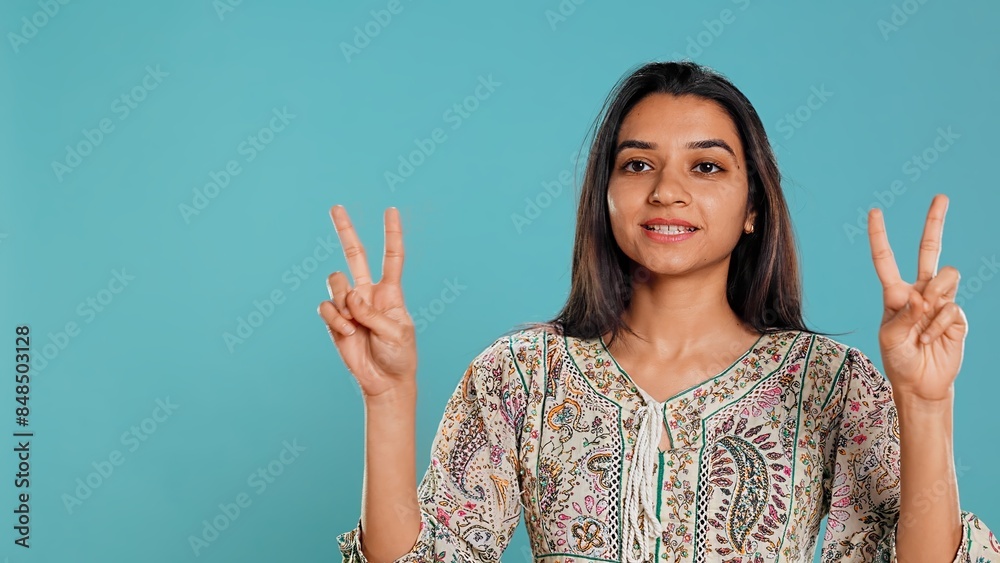 Jolly indian person doing peace victory hand sign gesture, studio backdrop. Portrait of upbeat woman, feeling confident, celebrating win, isolated over studio background, camera B Jolly indian person doing peace victory hand sign gesture, studio backdrop. Portrait of upbeat woman, feeling confident, celebrating win, isolated over studio background, camera B
