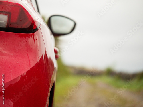 A red car is parked on a small country road with a view of stone fence and the sky. The car is wet from rain. Concept of calm and tranquility. Travel and tourism in Ireland concept. Selective focus