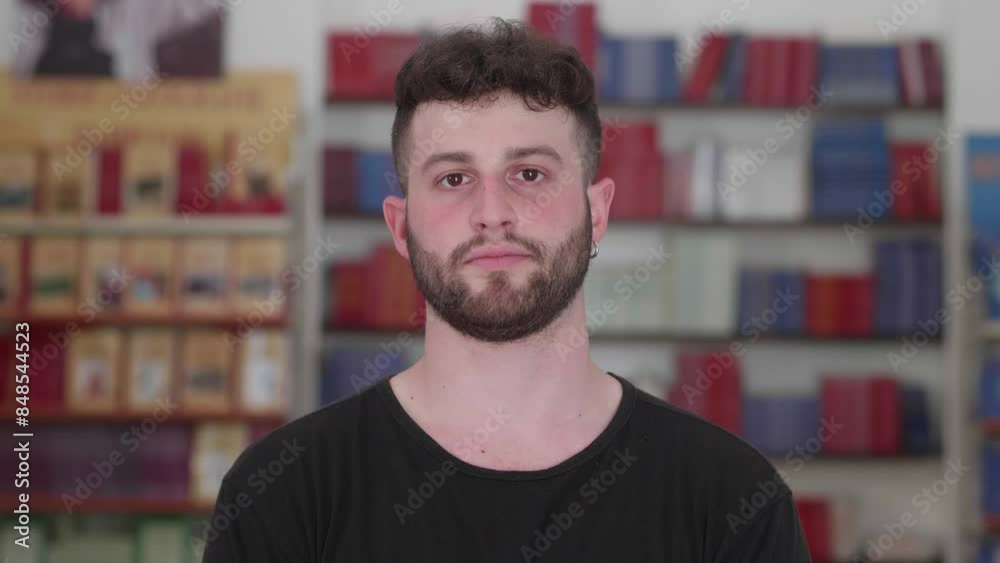 A young man with short black hair and a neatly trimmed beard, wearing a black shirt, looks seriously at the camera while standing in a bookstore. His expression is thoughtful and intense. A young man with short black hair and a neatly trimmed beard, wearing a black shirt, looks seriously at the camera while standing in a bookstore. His expression is thoughtful and intense.