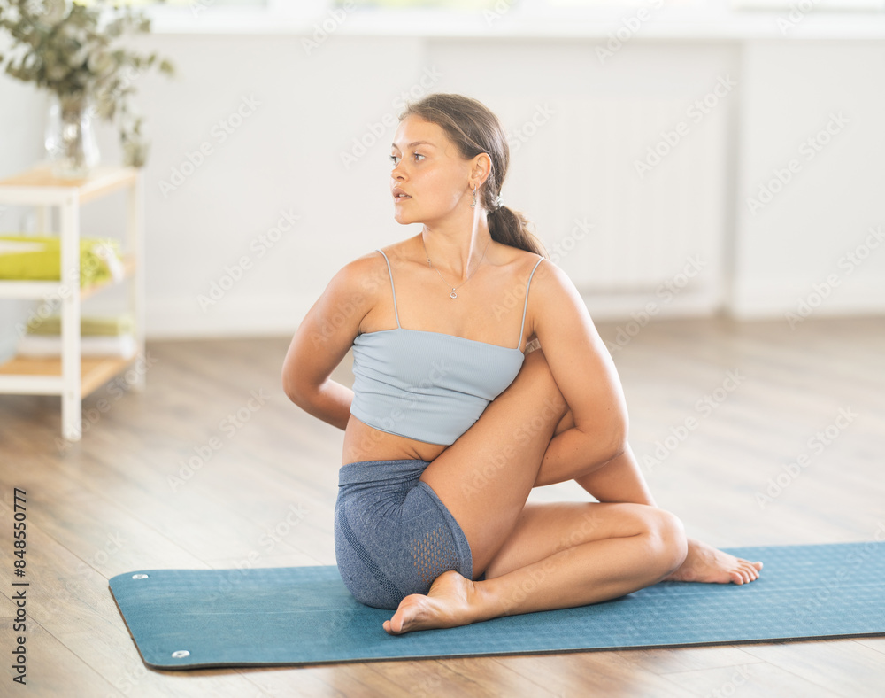 Obraz premium Sporty young girl performing seated twisting asana Matsyendrasana on mat in cozy sunlit room during daily yoga session..