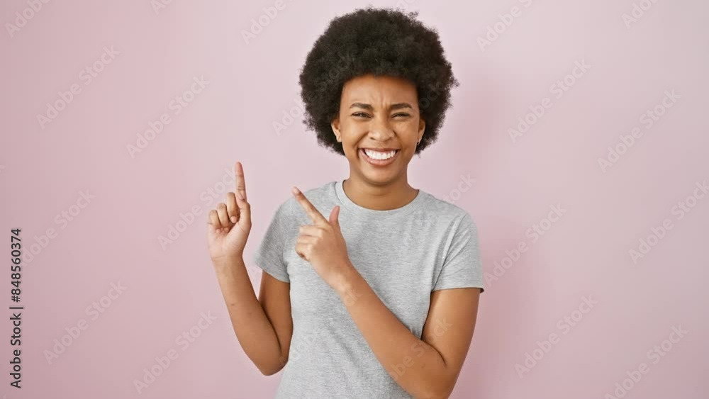 Cheerful african american woman in t-shirt, confidently pointing sideways with both hands, smiling at camera against isolated pink background