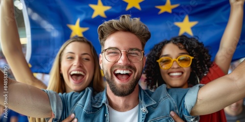 A group of three joyful people, two women and one man, celebrating together while holding a large European Union flag in a festive atmosphere photo