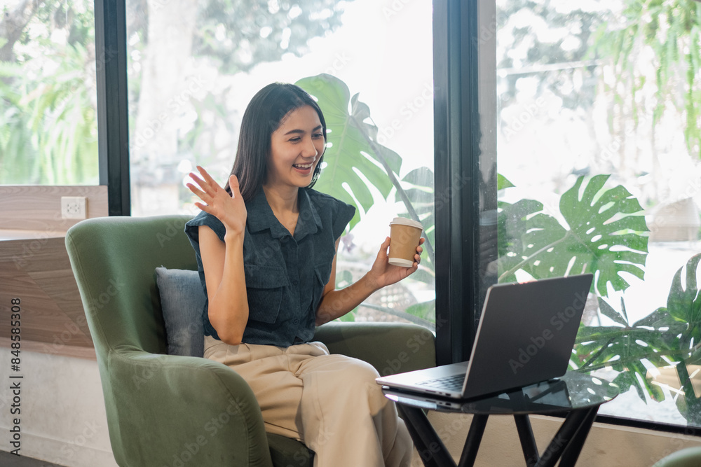 A cheerful young woman waves during a virtual meeting while holding a coffee cup, seated in a modern living room with large windows and lush greenery outside. A cheerful young woman waves during a virtual meeting while holding a coffee cup, seated in a modern living room with large windows and lush greenery outside.