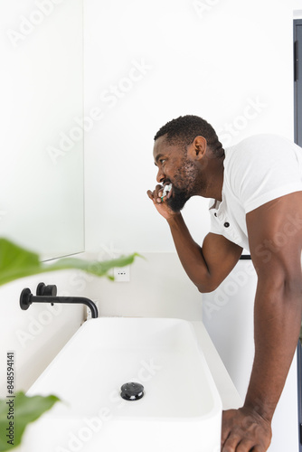 Young African American man brushing teeth in a modern bathroom with bright white walls, at home