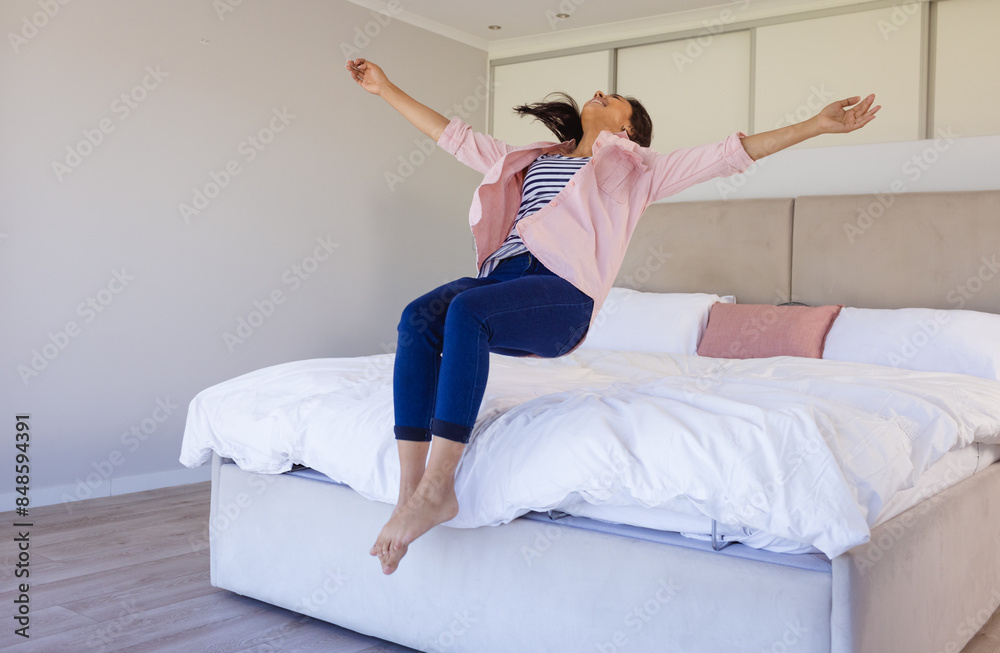 Happy biracial woman jumping on bed with arms outstretched, looking joyful