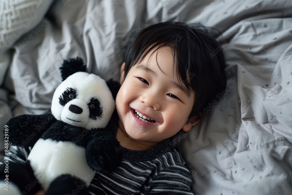 In the top view, an Asian child is lying on the bed, playing with a panda plush toy and smiling happily In the top view, an Asian child is lying on the bed, playing with a panda plush toy and smiling happily