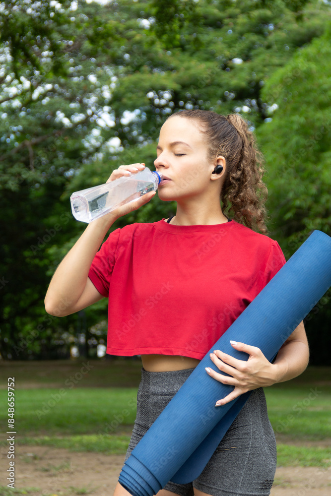 Beautiful latin hispanic female drinking water while holding yoga mat in the outdoors. Hydrating during workout stretching in a public park. Beautiful latin hispanic female drinking water while holding yoga mat in the outdoors. Hydrating during workout stretching in a public park.