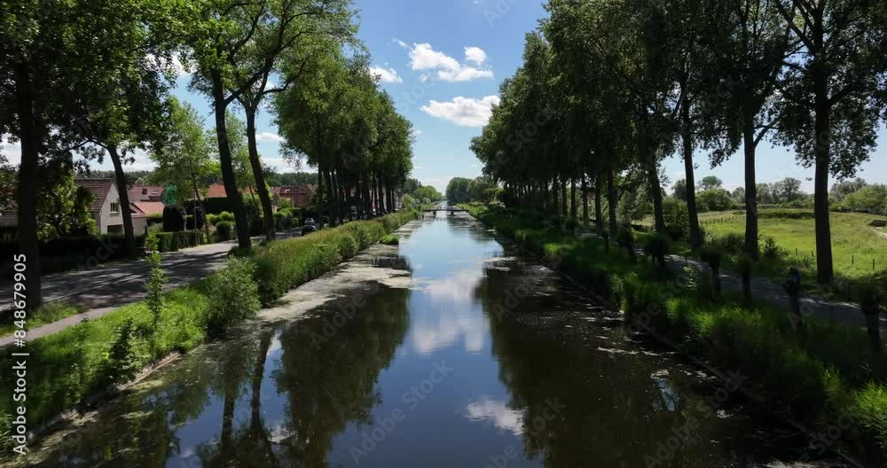 Damme, Belgium, June 2022. Drone pushes forwards above the Damse Vaart a peaceful canal lined with trees under a bright blue sky with fluffy clouds reflecting in calm water. Damme, Belgium, June 2022. Drone pushes forwards above the Damse Vaart a peaceful canal lined with trees under a bright blue sky with fluffy clouds reflecting in calm water.