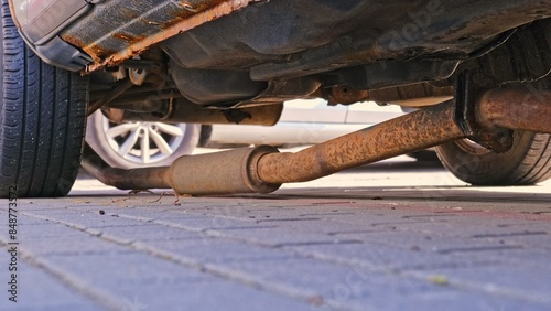Broken Exhaust Pipe Muffler Laying on Ground Under Old Car with Rusty Undercarriage Chassis