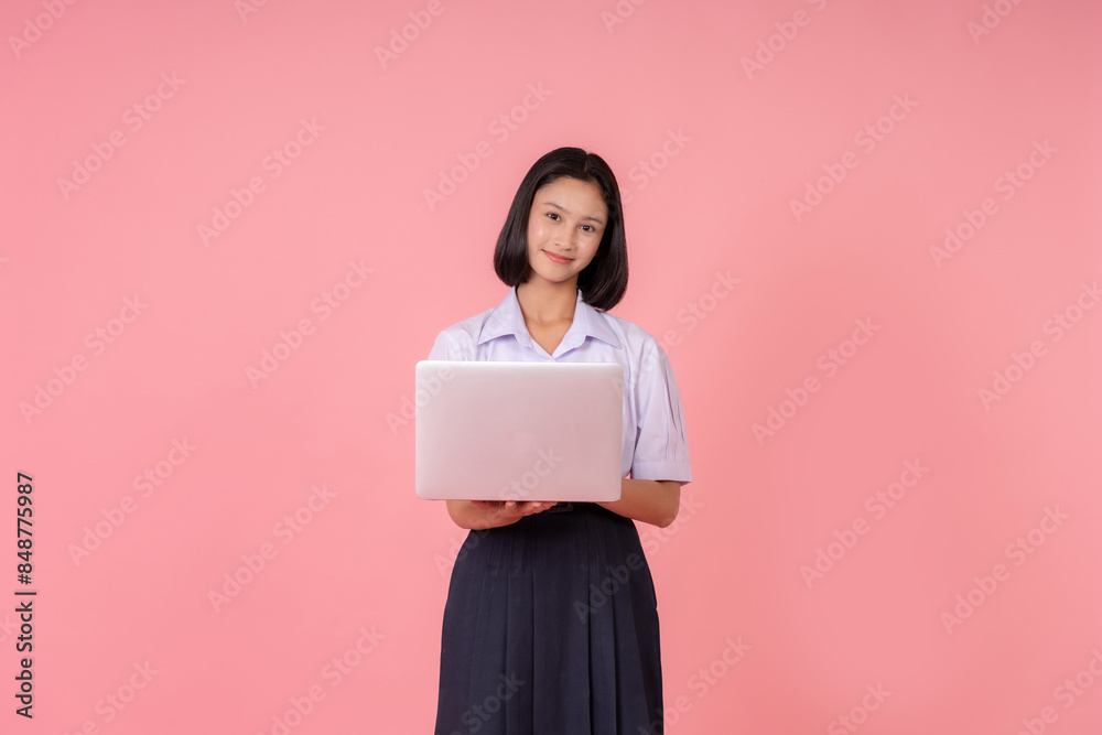 Half-body photograph of an Asian female high school student. Holding a laptop for reading, preparing for final exams and studying, posing for a photo in a studio with a pink background.