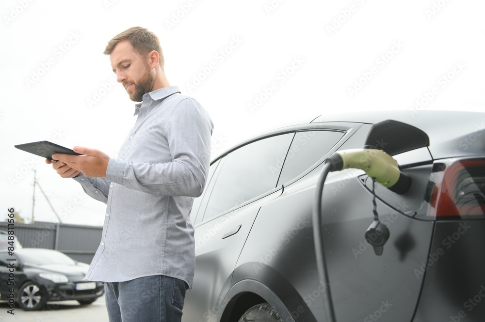 handsome bearded man in casual wear, standing at the charging station and charger for an electric car. Eco electric car concept handsome bearded man in casual wear, standing at the charging station and charger for an electric car. Eco electric car concept