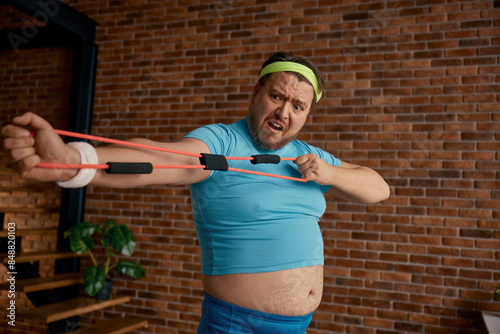 Obraz na plátne Comic chubby man in sportswear fooling around with fitness equipment