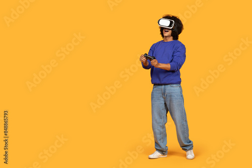 A man wearing a blue sweater and jeans stands in a yellow room, fully immersed in a virtual reality game. He holds a game controller and appears to be focused on his gaming experience.