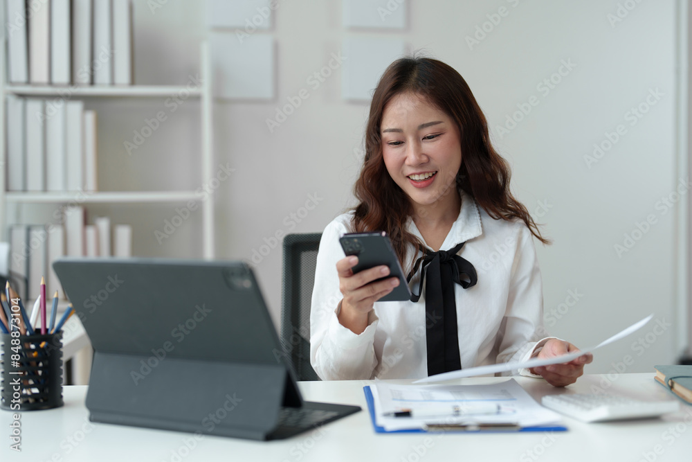 Happy Asian businesswoman in office while using smartphone to contact customer, online video call on documents, business finance, marketing, company taxes. Online business contact. Happy Asian businesswoman in office while using smartphone to contact customer, online video call on documents, business finance, marketing, company taxes. Online business contact.