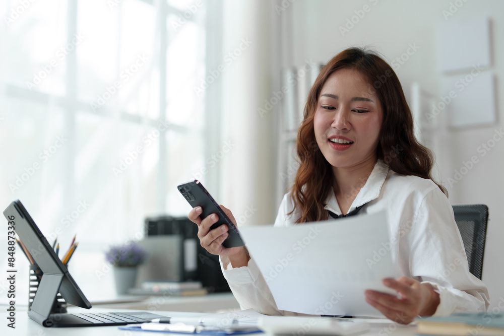 Happy Asian businesswoman in office while using smartphone to contact customer, online video call on documents, business finance, marketing, company taxes. Online business contact.