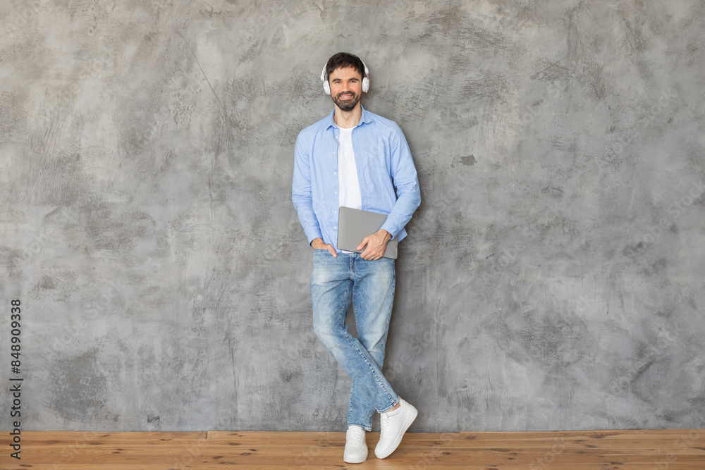Man standing in front of a concrete wall, leaning against it with one hand in his pocket. He is smiling at the camera and wearing headphones over his ears. He is holding a laptop in his other hand Man standing in front of a concrete wall, leaning against it with one hand in his pocket. He is smiling at the camera and wearing headphones over his ears. He is holding a laptop in his other hand