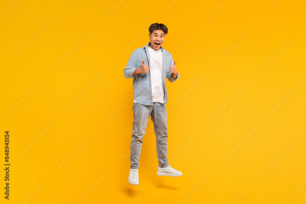 This image shows a young man in a blue shirt and jeans jumping in front of a yellow background. He is smiling and giving a thumbs up with both hands. He seems to be happy and excited about something. This image shows a young man in a blue shirt and jeans jumping in front of a yellow background. He is smiling and giving a thumbs up with both hands. He seems to be happy and excited about something.